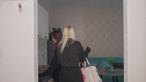young woman hands jug to friend on green couch in stylish apartment kitchen while other fills jug with water from sleek chrome tap, capturing warm supportive interaction and modern home atmosphere