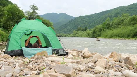 a male traveler is resting lying in a green tent he uses a smartphone to communicate a male traveler is resting lying in a green tent he uses a smartphone to communicate concept of resting in the mountains