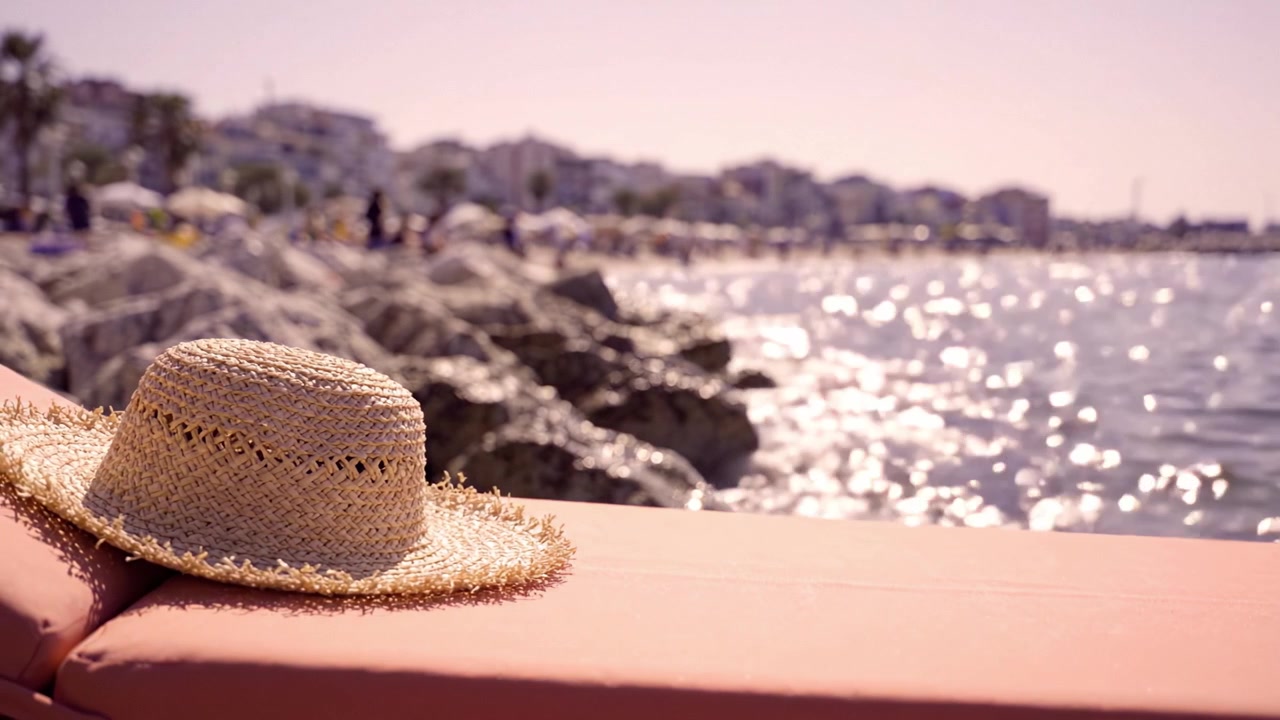 Una splendida giornata in spiaggia: un cappello di paglia appoggiato su una ringhiera sul mare con acqua scintillante e un paesaggio illuminato dal sole sullo sfondo, ideale per l'atmosfera estiva.