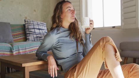 thoughtful caucasian woman sitting on floor in living room drinking coffee, looking out of window