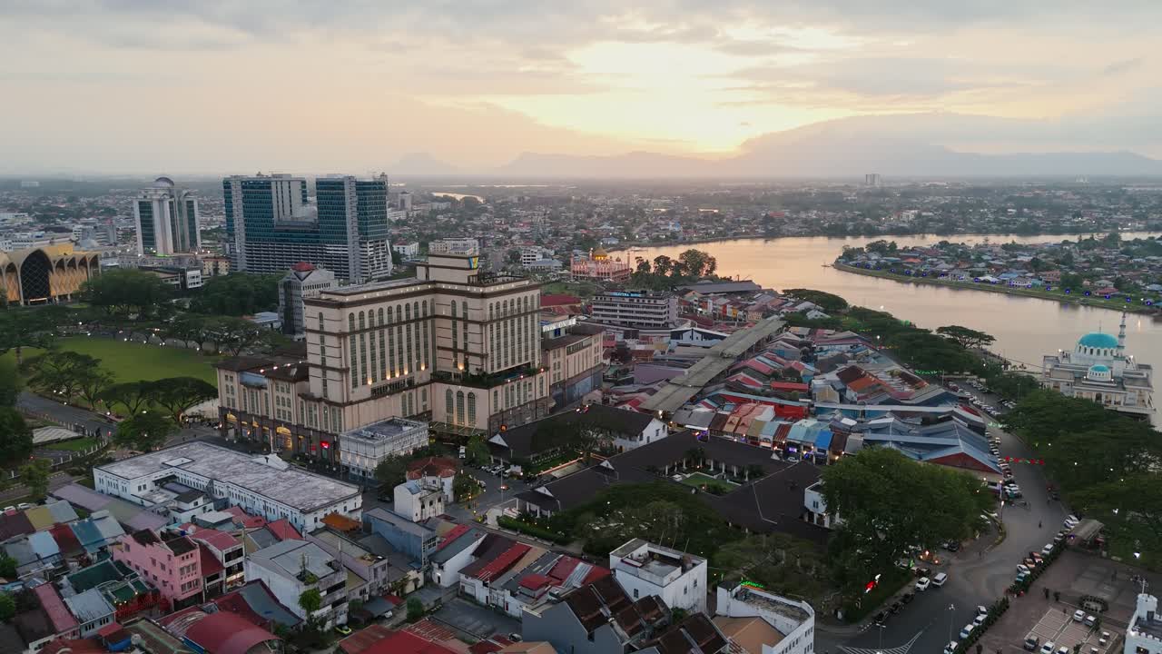 Golden hour drone shot of Kuching’s old town, lined with traditional shop houses and framed by the Sarawak River, with distant hills and the setting sun painting the scene warmly.