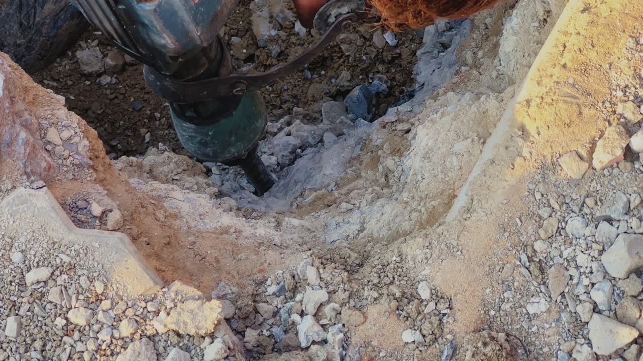 Close-up view of drilling machine bit penetrating soil and rock inside dug pit, rough textures and loose earth visible in a construction or excavation outdoor setting