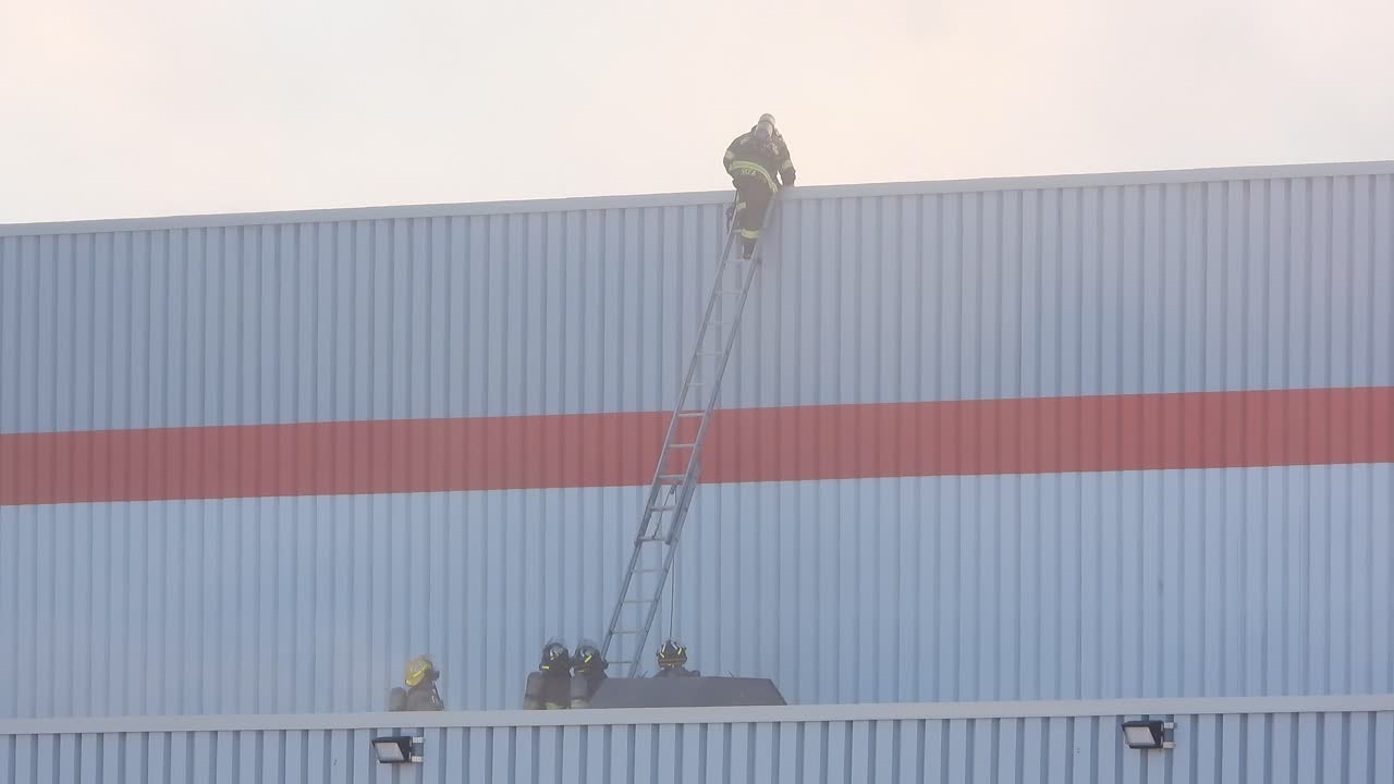 Firefighter walks towards edge and climbs down ladder up against metal building