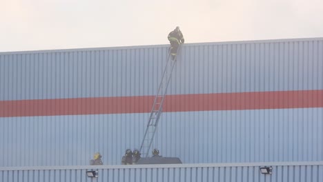 Firefighter-walks-towards-edge-and-climbs-down-ladder-up-against-metal-building