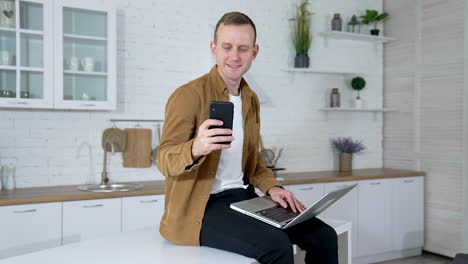 portrait of a student with a laptop and a phone. smiling young man is sitting on a kitchen table and using a laptop and mobile phone at home. quarantine concept.