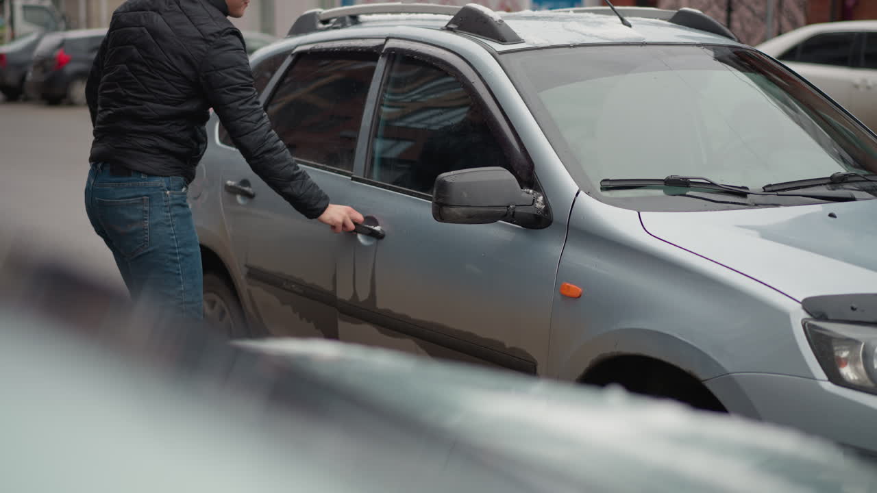 Urban Man wearing black winter coat and blue jeans opens and closes car door then enters ash coloured vehicle parked on urban street with water droplets and snow on surface in cold winter scene