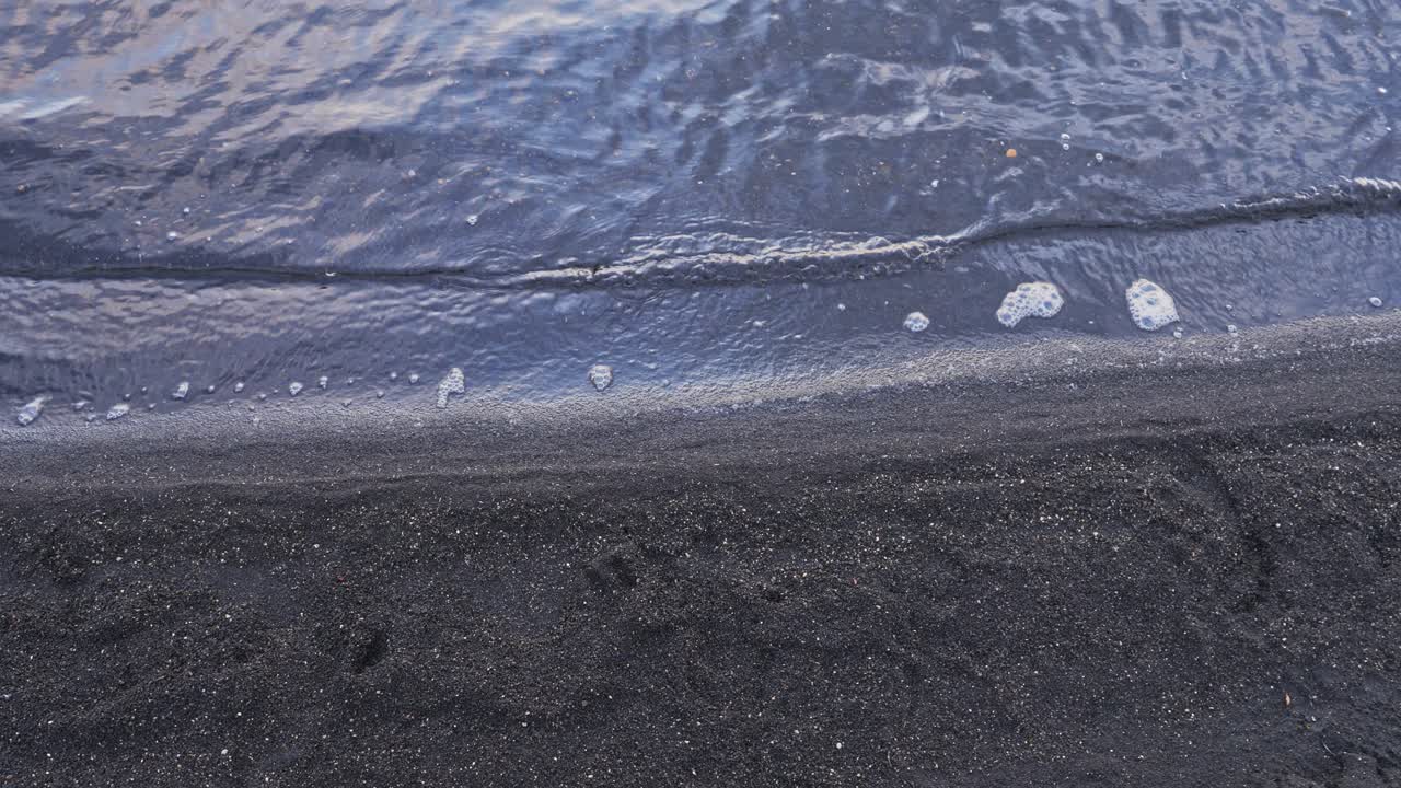 Close-up, Handheld shot of small waves washing up onto a dark, black sand or volcanic ash beach