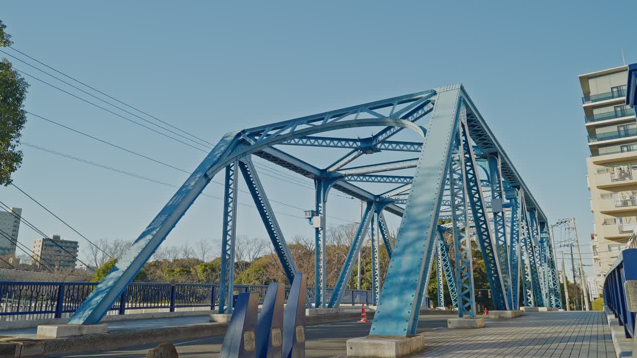 Blue steel truss bridge spans across a clear sky in Tokyo, with residential buildings and bare trees visible alongside, captured as a static low-angle shot emphasizing geometric metalwork