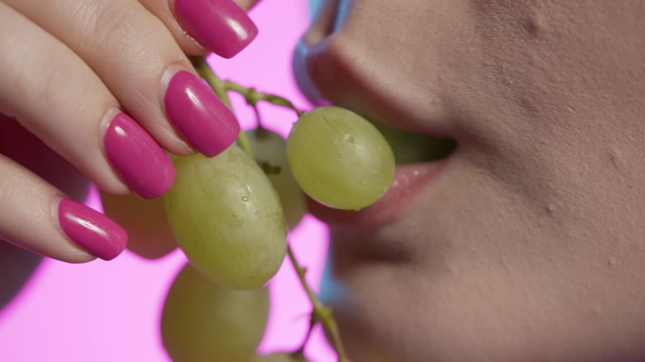 mujer comiendo uvas blancas en primer plano, esmalte de uñas femenino y labios de lápiz labial rojo en fondo rosa