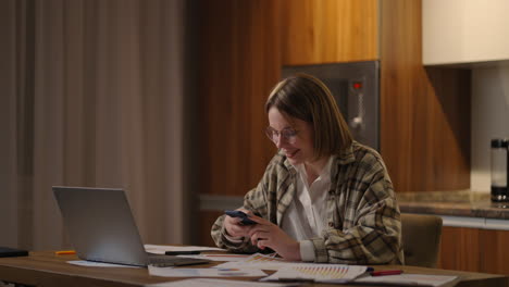 Glückliche-Frau,-Die-Ihr-Mobiltelefon-Benutzt,-Während-Sie-Zu-Hause-Mit-Einem-Laptop-Arbeitet.-Lächelnde,-Coole-Frau-Mit-Brille,-Die-Nachrichten-Mit-Dem-Smartphone-Sendet.-Schöne,-Stilvolle-Dame,-Die-Auf-Dem-Handy-Auf-Der-Website-Surft