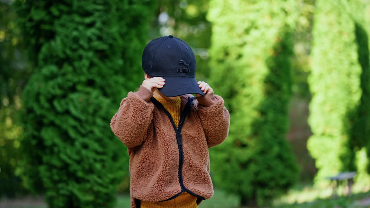 Cute baby boy in brown jacket holds a black cap. Kid puts on the cap hiding behind it. Greenery at backdrop.