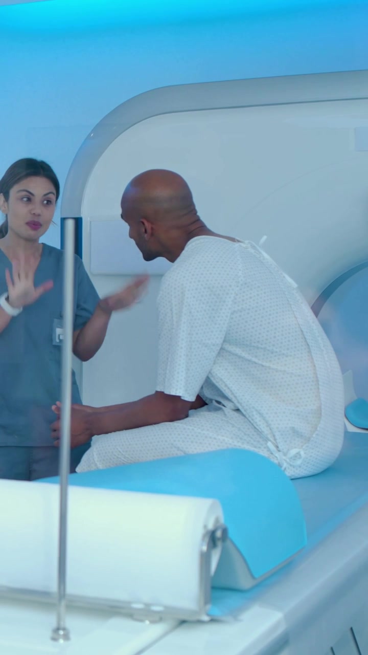 Vertical video: Tech in scrubs gesturing, patient leaning on MRI bed in gown for scan, copy space