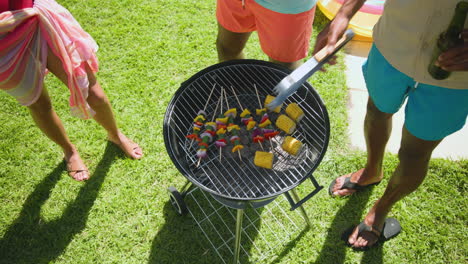 diverse friends enjoying barbecue by pool, grilling skewers and corn, holding drinks