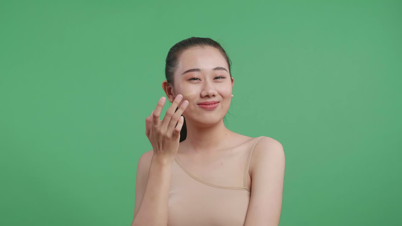 미소 짓는 아시아 여성이 스튜디오에서 녹색 스크린 배경에 서서 얼굴과 어깨에 크림을 적용합니다.