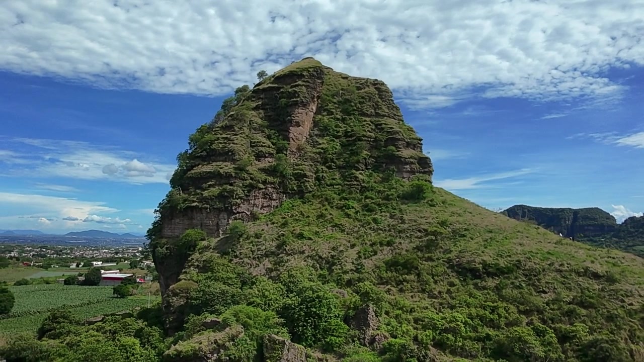Imagem aérea de drone da colina rochosa Cerro Hotcakes coberta por vegetação verde exuberante sob um céu azul com nuvens texturizadas, exibindo uma paisagem natural dramática durante o dia