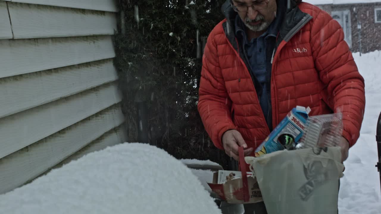 A man with arms loaded with garbage and recycling carries it out to trash bins around the side of the house as snow gently falls