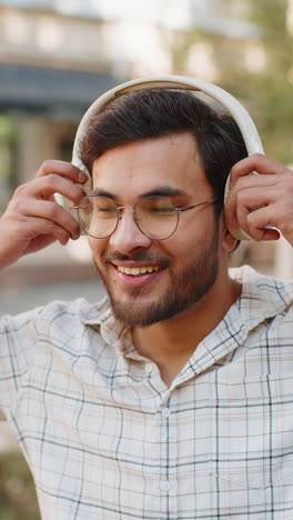 portrait of happy young indian man wearing wireless headphones listening to music on city street