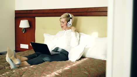 happy businesswoman in headphones listening music smiling dancing and singing whilewhile using laptop lying in bed at hotel room. travel, business and people concept