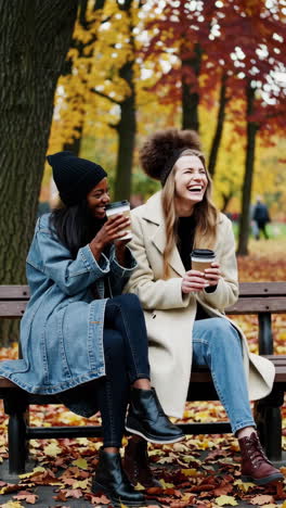 two women laughing and drinking coffee on a park bench in autumn