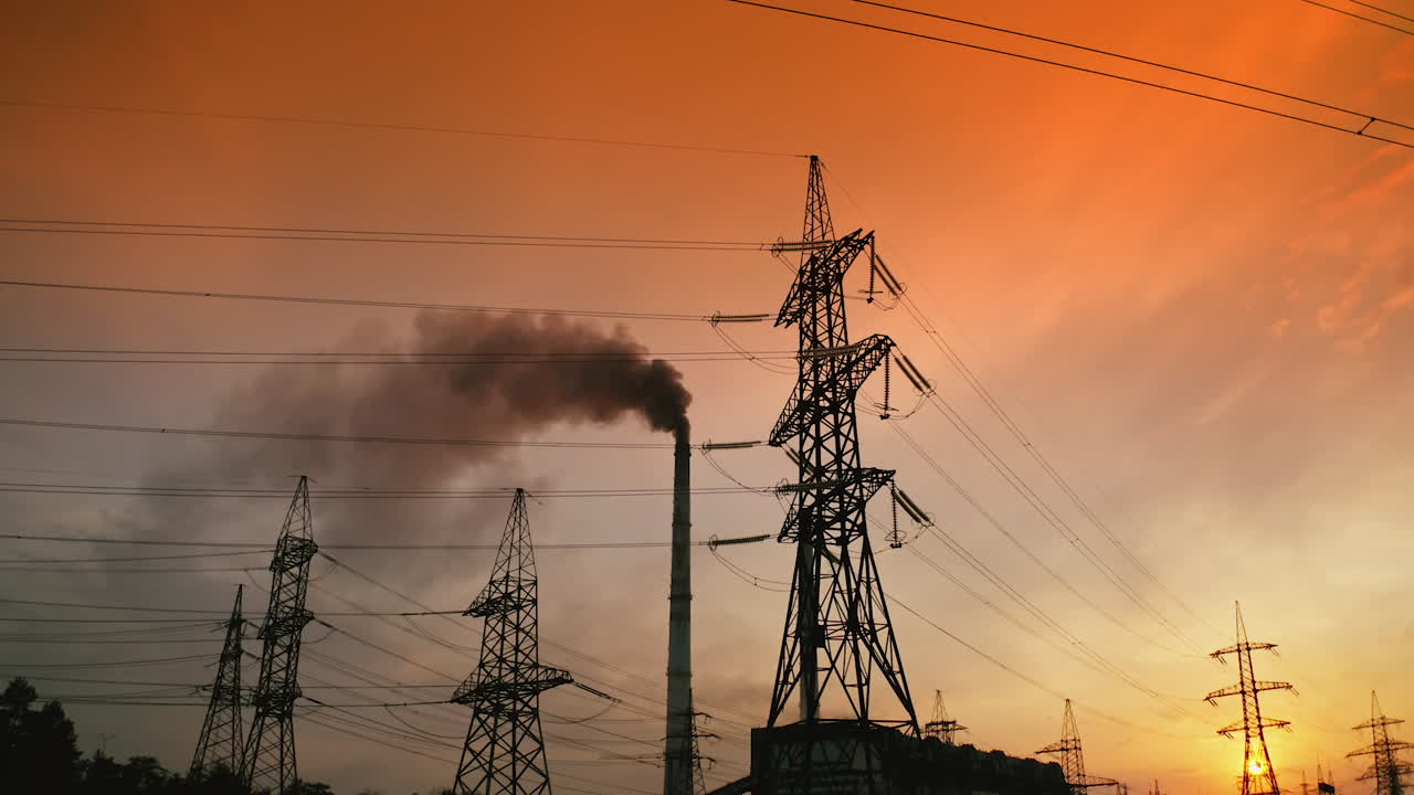 Transmission lines near smoking industrial pipe at sunset. High-voltage electric power line and dark smoke released from factory in the evening.