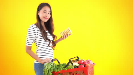 Mujer-Delgada-Leyendo-Una-Etiqueta-De-Un-Bocadillo-Mientras-Compra-Comida-Y-Verduras-En-Un-Fondo-Amarillo-Aislado