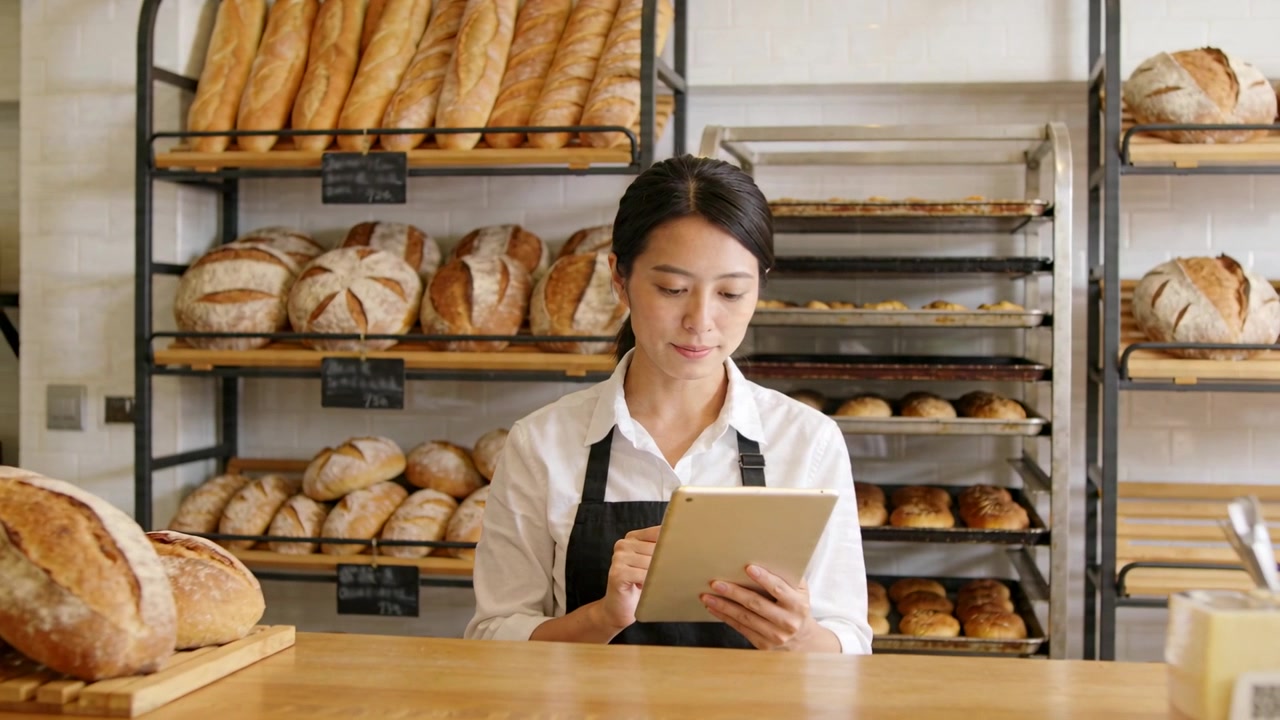 Fornaio con camicia bianca e grembiule nero che tocca il tablet dopo un segnale acustico, controllando gli ordini al bancone con una pagnotta