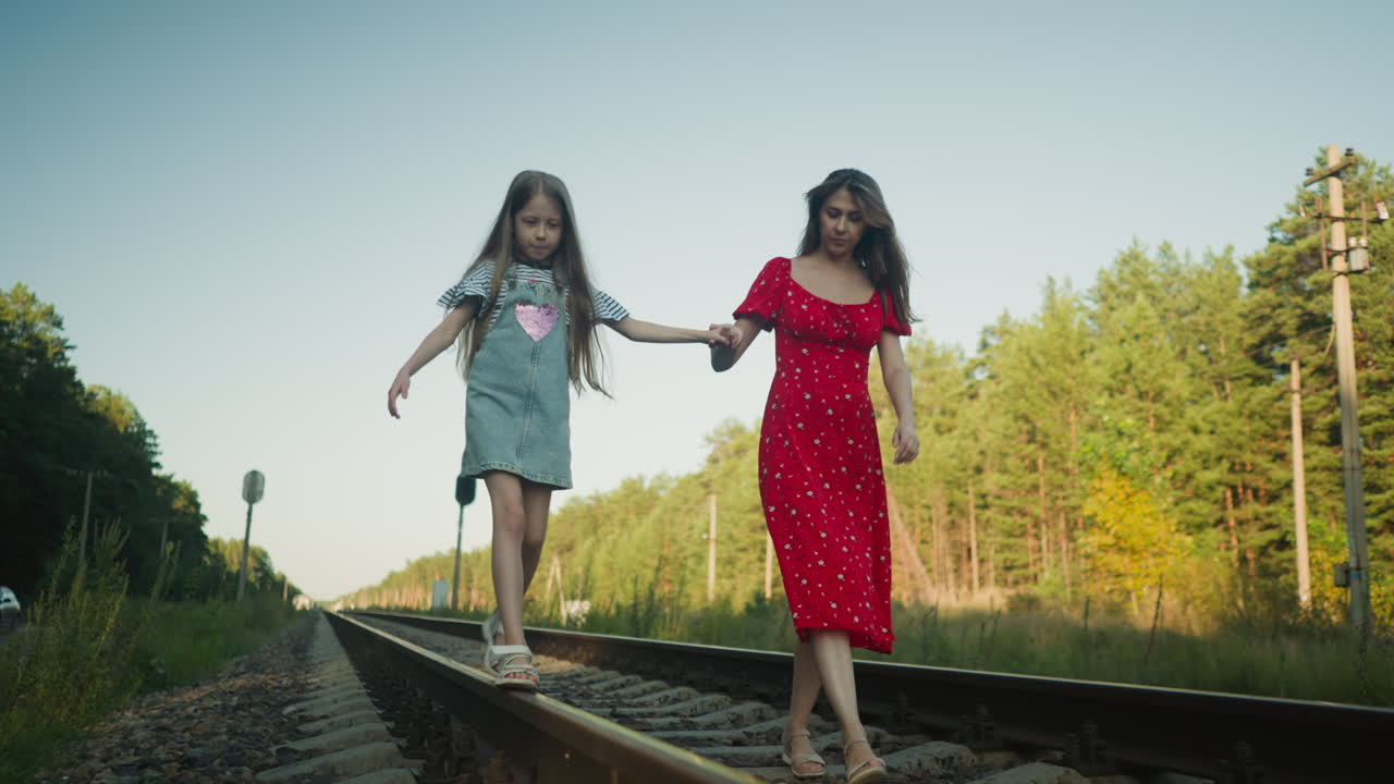 woman gently holding daughter hand as she balances on rail beam, surrounded by tall trees and wild greenery in peaceful countryside setting, with parked car subtly visible in distant background