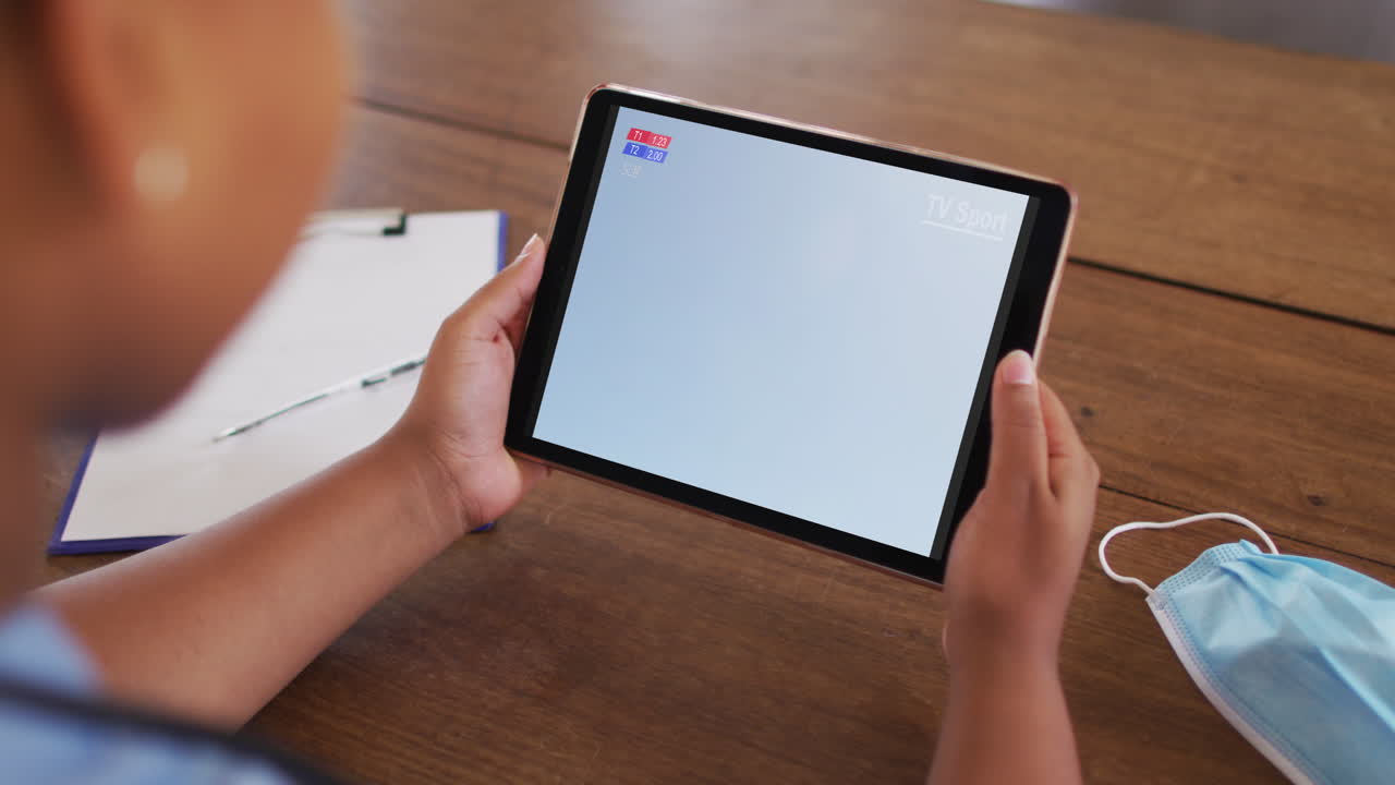 태블릿에서 축구 경기를 시청하는 얼굴 마스크와 함께 테이블에 앉아있는 여성의 복합