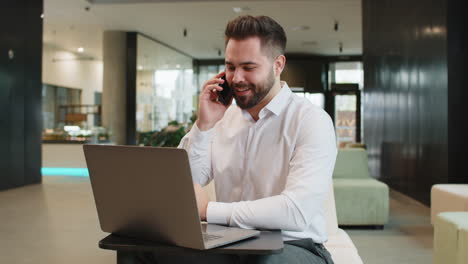 Businessman in formal shirt answering to client on mobile phone call using laptop in office lobby
