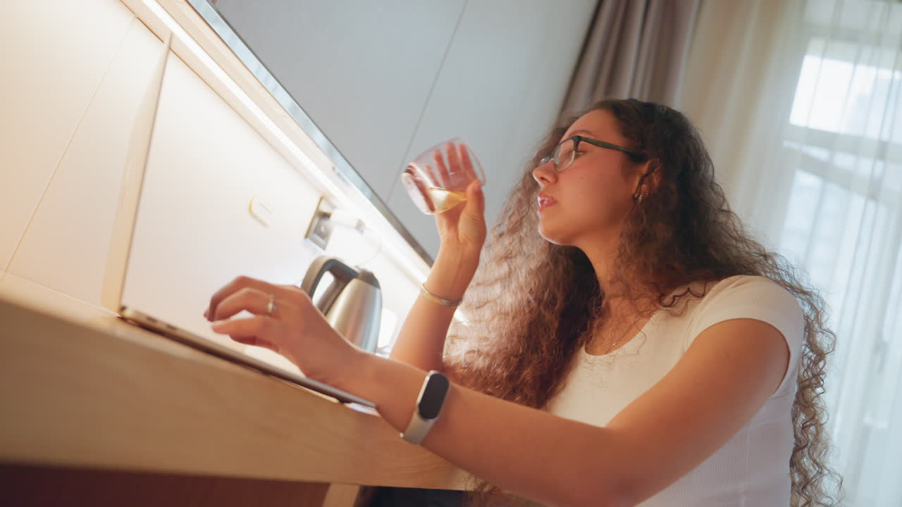 Ospite donna che lavora alla scrivania dell'hotel, donna dai capelli ricci che digita e sorseggia acqua, bollitore sulla scrivania, luce solare naturale attraverso le tende, lavoro da remoto e studio rilassati, atmosfera da scrittrice di viaggi, calda ospitalità