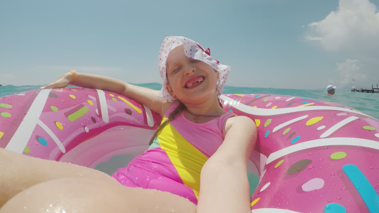 A Little Girl In A Cool Hat And A Bright Bikini Is Riding On A Circle And Making A Selfie Smiling At