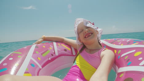 a little girl in a cool hat and a bright bikini is riding on a circle and making a selfie smiling at