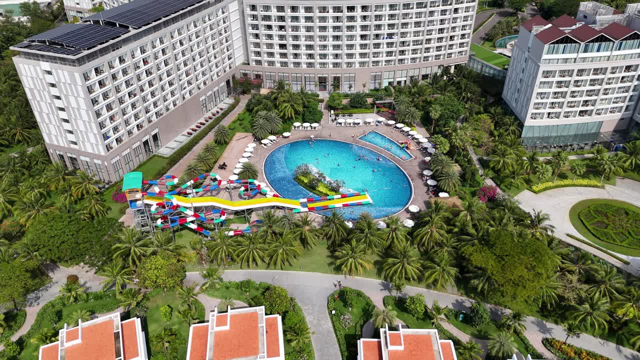 Late afternoon aerial footage of a resort with large modern buildings around a central pool featuring water slides and lounge chairs, amidst lush tropical greenery, a family-friendly destination.