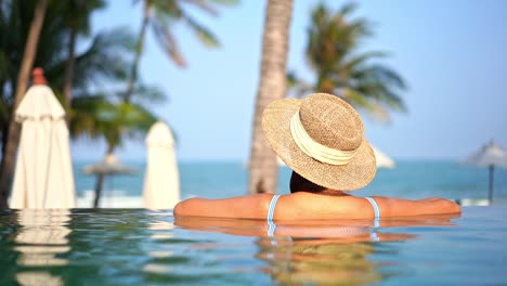 Rückseite-Der-Frau-Mit-Sommerhut-Im-Swimmingpool-Des-Luxusresots-Mit-Blick-Auf-Das-Offene-Meer-Und-Die-Tropische-Vegetation,-Vollbild