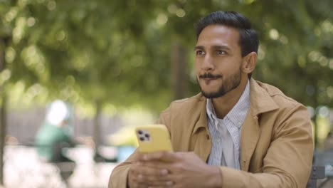 muslim man sitting at outdoor table on city street sending text message to date on mobile phone