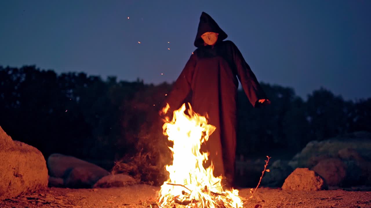 Mysterious witch near fire. Scary wizard in black costume with hood standing at fire with stretched arms. Halloween.