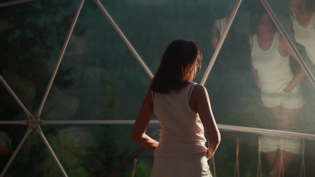 밤에는 거울 창문으로 바라보는 여성, 밤에는 고급스러운 친환경 캠프에서 휴가를 보내는 관광객, 유리 벽에 반사된 여성.