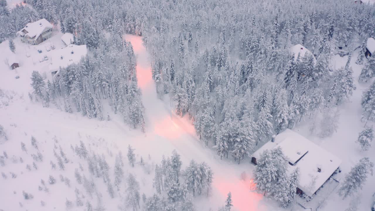 Loittonevat ja alaspäin kallistuvat droonikuvat lumisesta mäntymetsästä, teistä ja mökeistä Suomen taiga-tunturi-alueella.