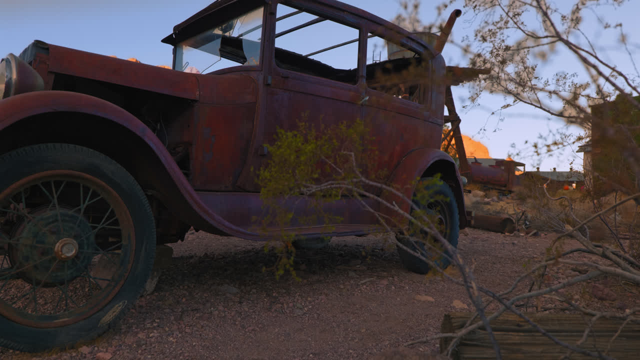 An old beat up, rusty truck sits with broken windshield in a desert. The camera dollies slightly from behind a bush to reveal the vintage vehicle.