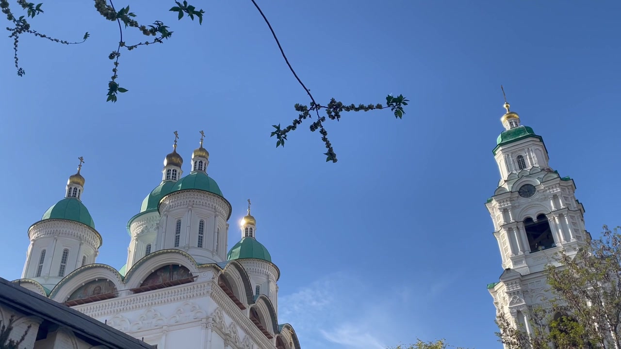 Vista esterna della Cattedrale dell'Assunzione nel Cremlino di Astrakhan, Russia, che mostra le mura in pietra bianca e le cupole verdi incorniciate da rami d'albero contro un cielo sereno.