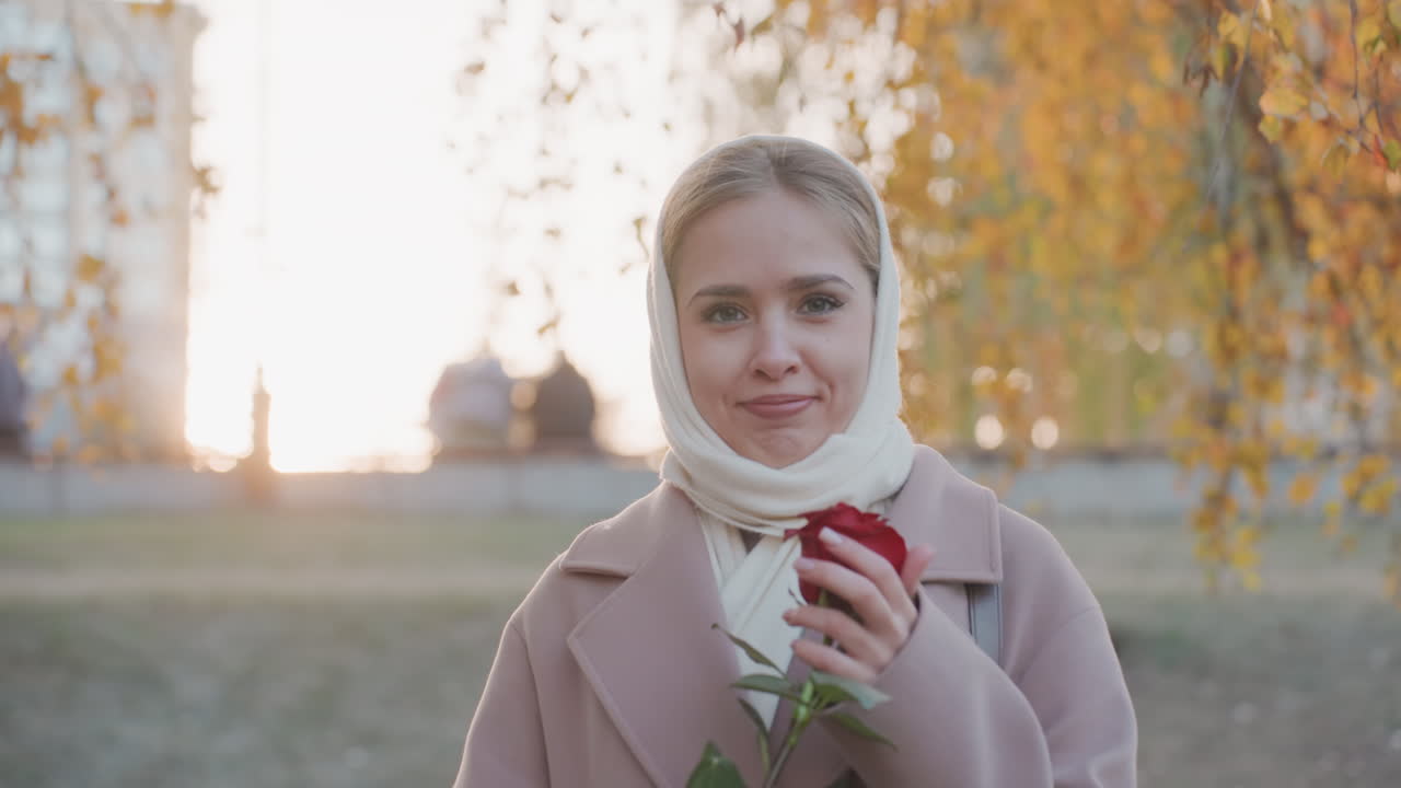 Mawar merah dan senyum hangat. Wanita nyaman dengan bunga merah di taman. Wanita tersenyum dengan mawar saat musim gugur di tengah pemandangan kota. Pemandangan perkotaan menampilkan wanita ceria memegang mawar selama golden hour di taman.
