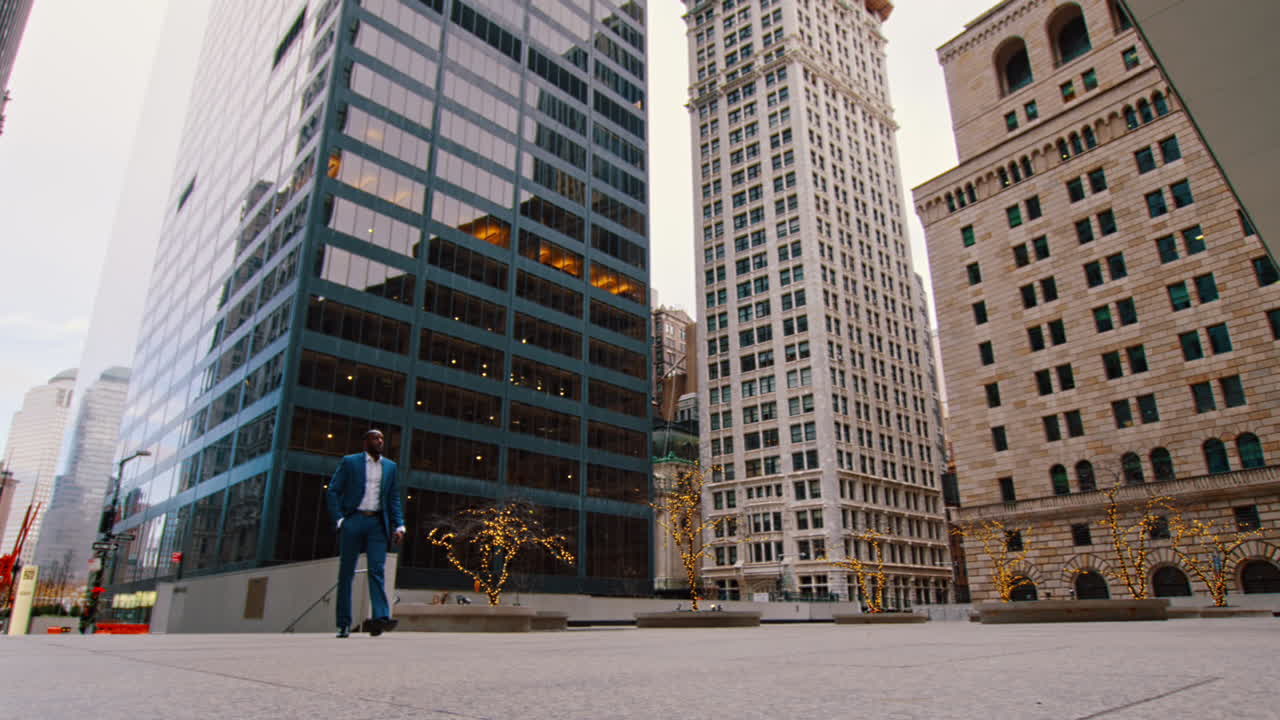Nova Iorque, EUA, 1 de abril de 2026: Empresário caminhando por uma praça de Manhattan perto de edifícios de escritórios. Imagem ampla de empresário negro de terno azul caminhando por uma praça urbana com torres de escritórios de Nova Iorque