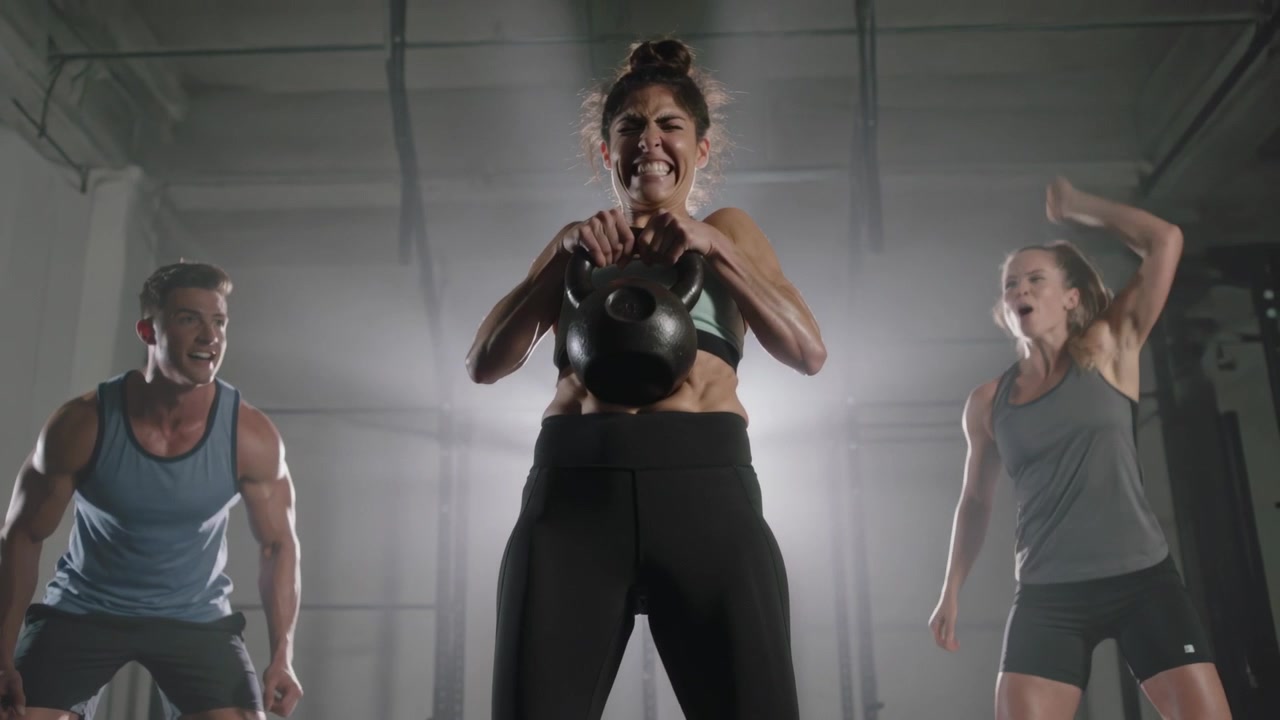 Un'atleta in reggiseno sportivo afferra un kettlebell e lo fa oscillare al petto in palestra, mentre i compagni di allenamento fanno il tifo.