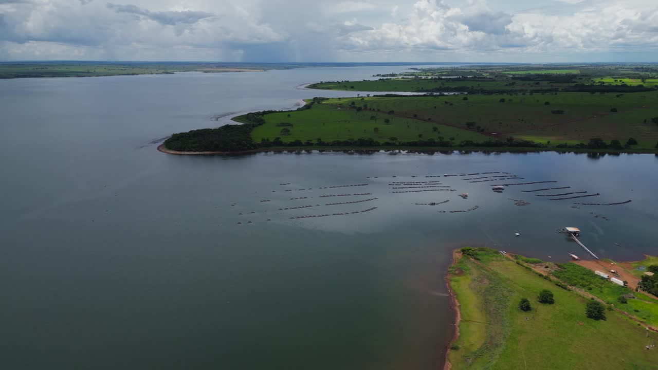 Aerial view of a large Brazilian reservoir featuring distinct aquaculture or fish farming structures in the water, bordering expansive green fields and a small pier area under a cloudy sky