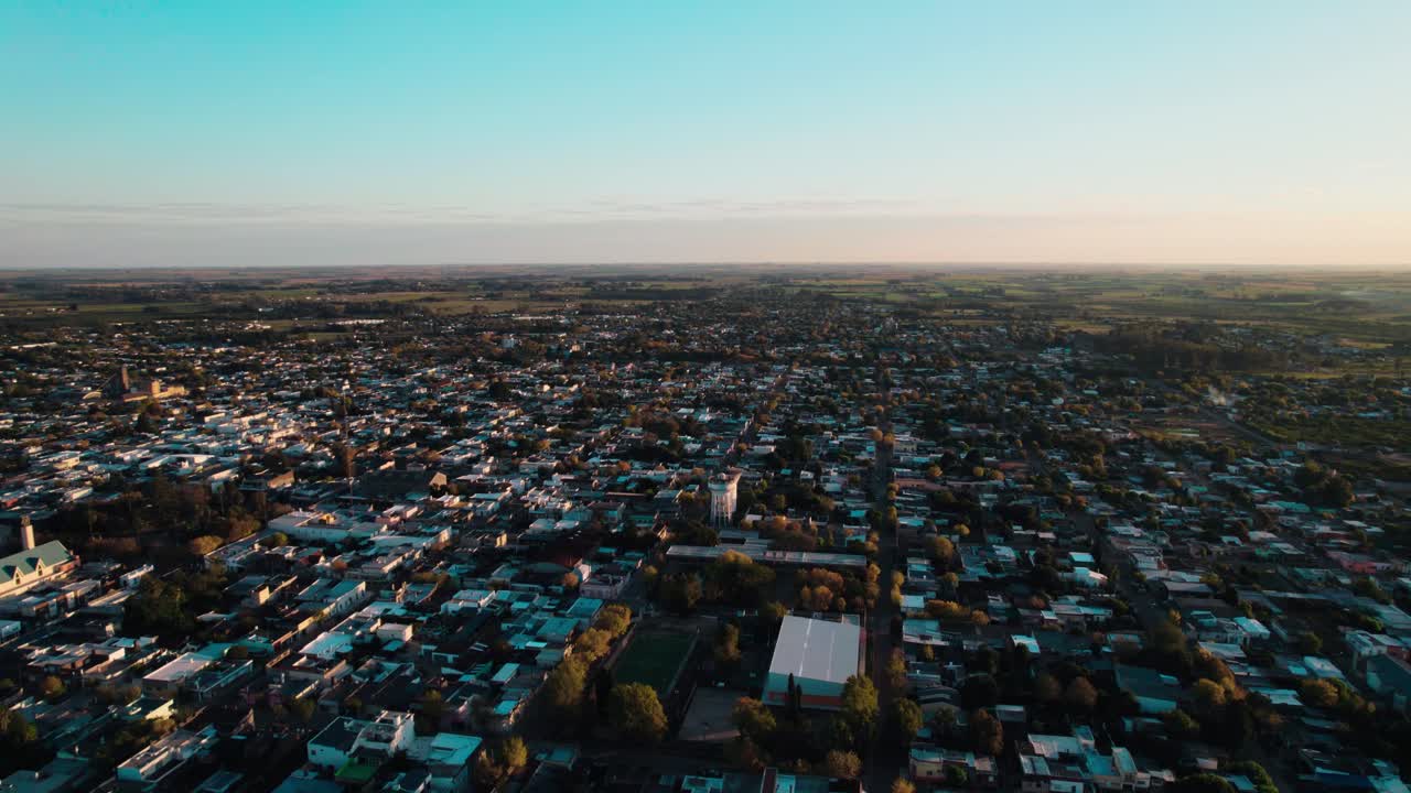 Drone shot capturing the full breadth of Carmelo, Uruguay, with low-rise buildings and tree canopy extending into the horizon under a golden hour sky. Features a peaceful, wide view of the town