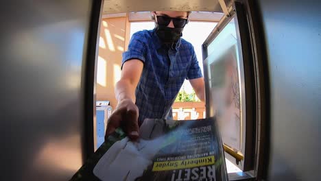 pov from inside of a newspaper vending machine, a man wearing a mask and earbuds opens the container removing a magazine from inside