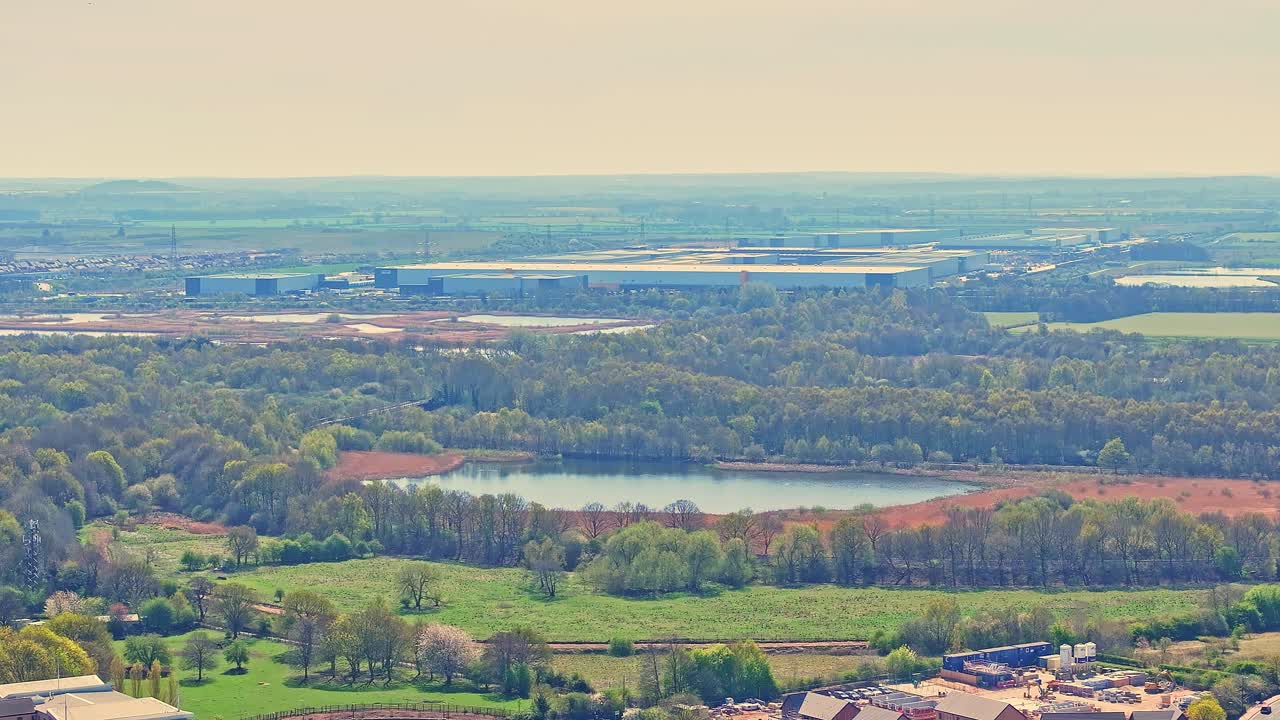 Drone captures Doncaster Lakeside area featuring dense forest, wetlands with small lake, industrial warehouses, and open green fields under hazy sky in South Yorkshire, UK