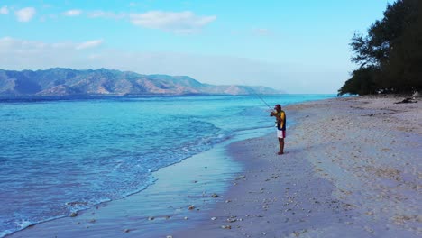 Angelbegeisterter-Junger-Mann,-Der-Versucht,-Fischköder-Zu-Reparieren-Und-Angelrute-Vom-Strand-Zum-Ruhigen-Blauen-Meer-An-Der-Balinesischen-Küste-Zu-Werfen