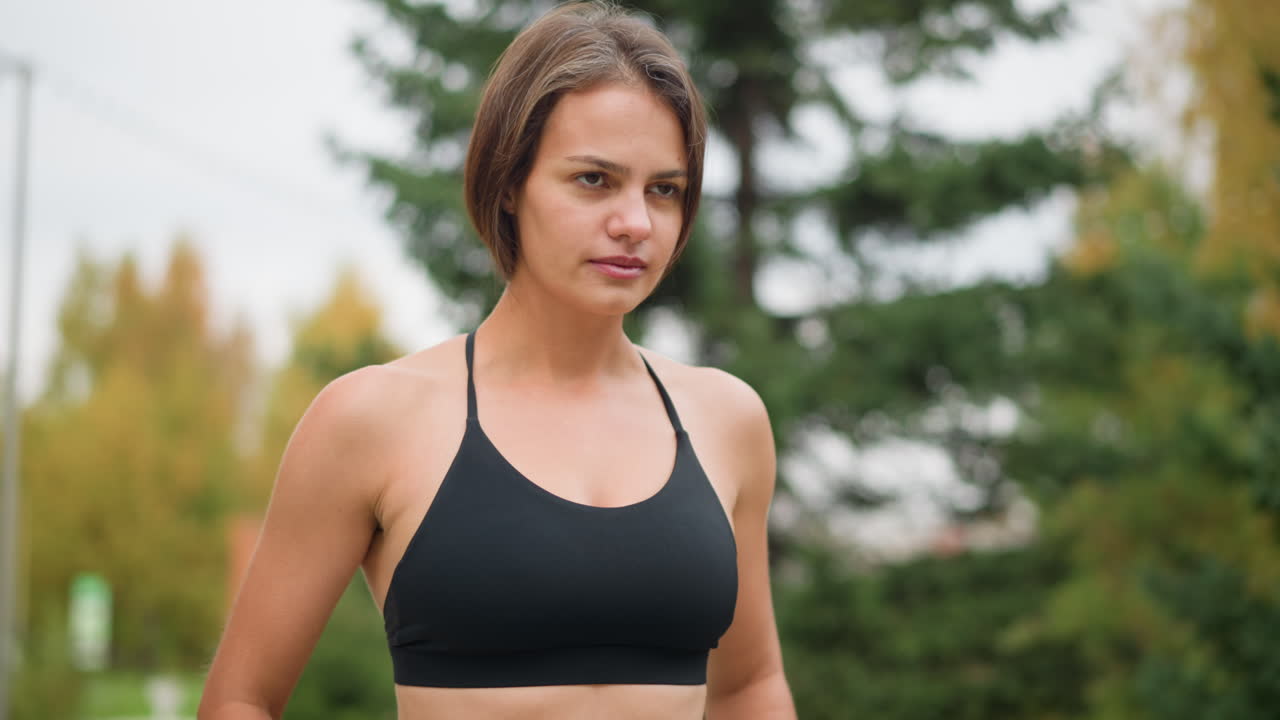 Close-up view of girl in neck top breathing in deeply and putting hand on waist, relaxed pose, calm expression, outdoor setting, deep breath, focus moment, fitness and health, natural shot