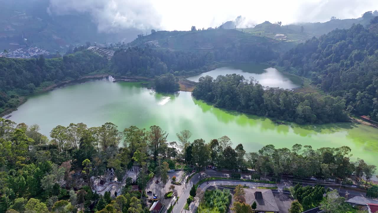 Drone view of serene highland lake with vibrant hues of green and blue, surrounded by dense forest and mist-covered mountains. Peaceful and picturesque natural scenery, Telaga warna, Dieng, Indonesia
